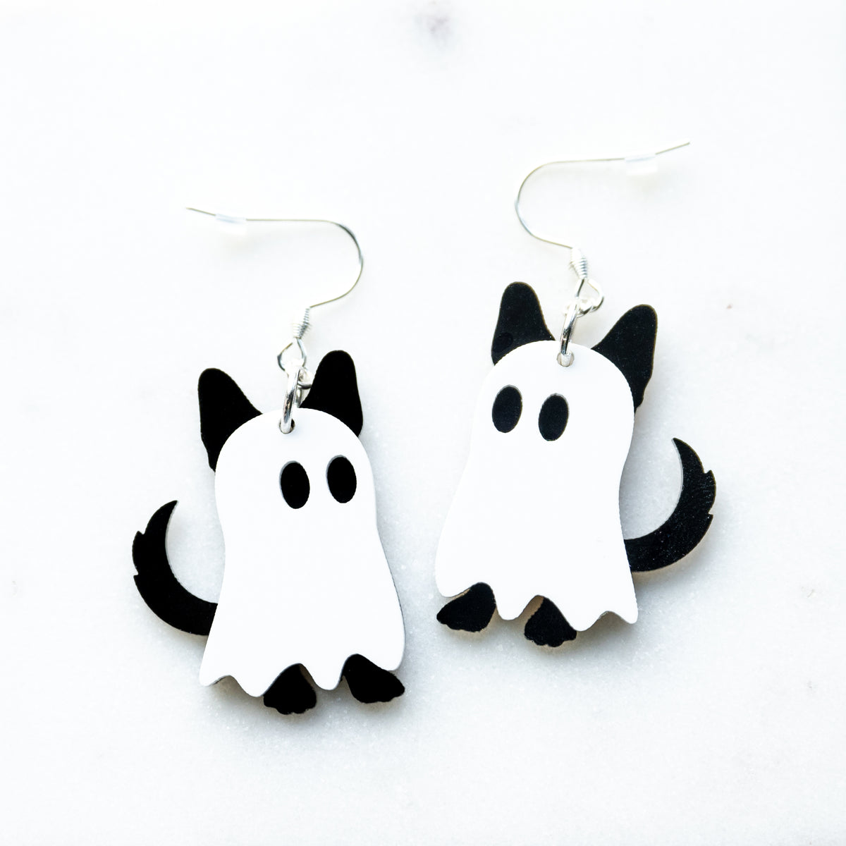A pair of white and black ghost-shaped dangle earrings with dog ears, feet and tail, designed to look like ghosts dogs with fish hook hardware on a white background.