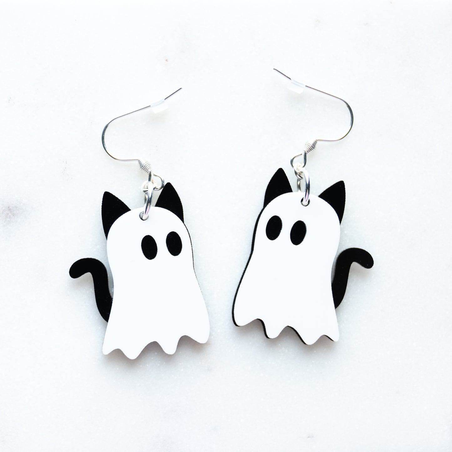 A pair of white and black ghost-shaped dangle earrings with cat ears and tail, designed to look like cat ghosts with fish hook hardware on a white background.