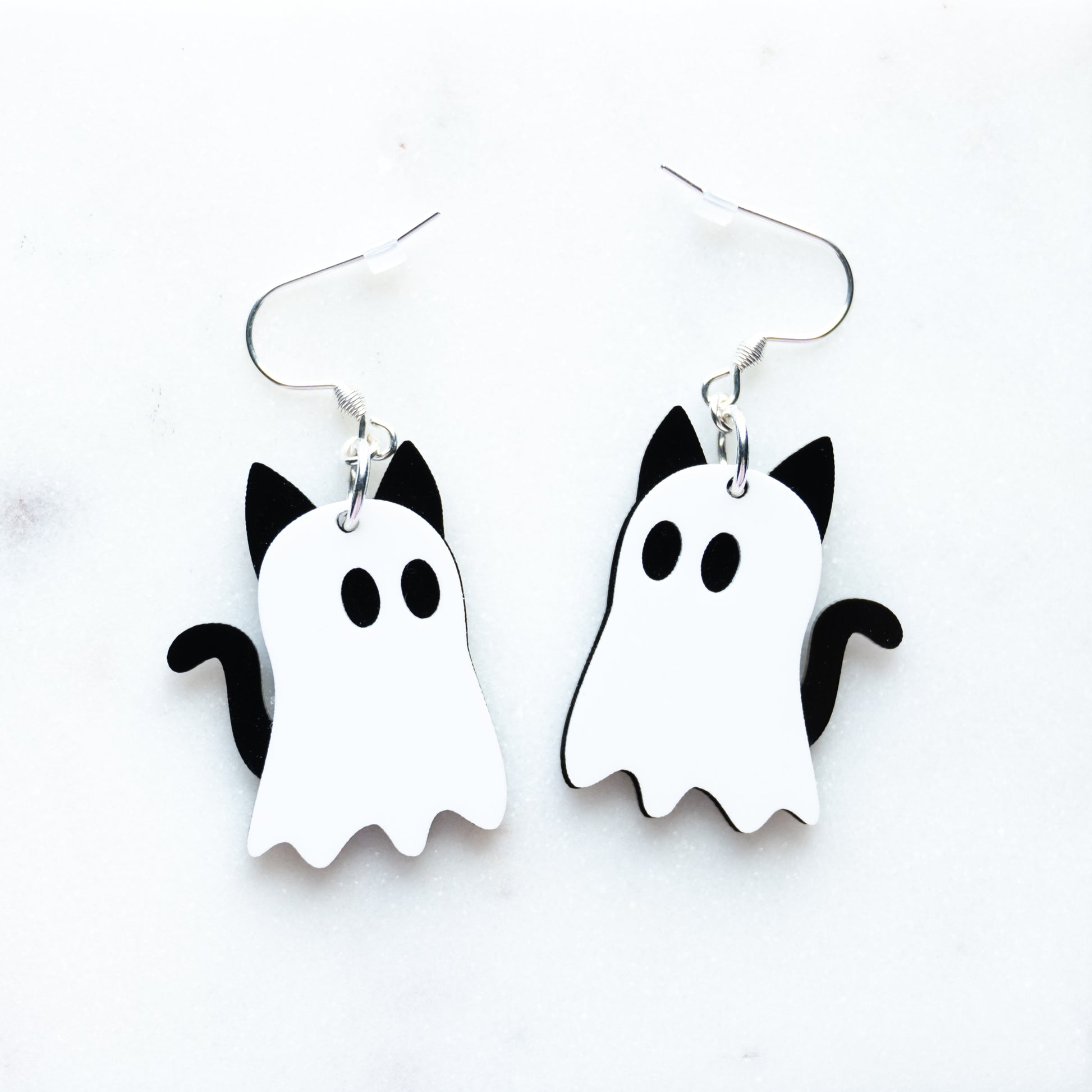 A pair of white and black ghost-shaped dangle earrings with cat ears and tail, designed to look like cat ghosts with fish hook hardware on a white background.
