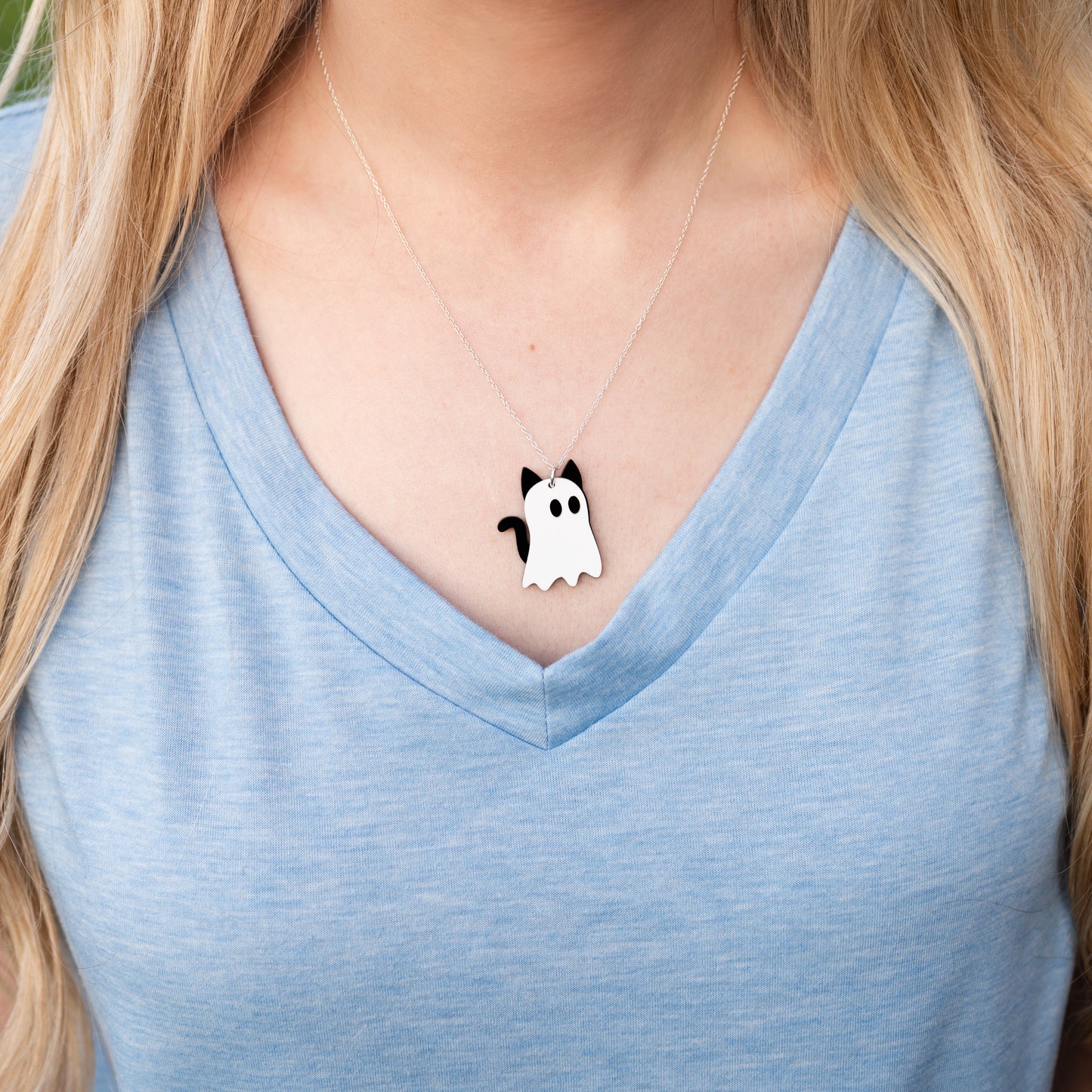 A white and black ghost-shaped acrylic necklace with cat ears and tail, designed to look like cat ghosts with a cable chain on a model with long blonde hair wearing a light blue v-neck shirt. 