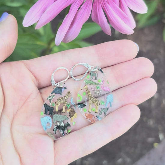 Video showing teardrop shaped acrylic earrings with a zoo animal pattern, including representations of various animals such as a giraffe, lion, zebra, and others, displayed on a hand with a grassy background with a pink flower. 