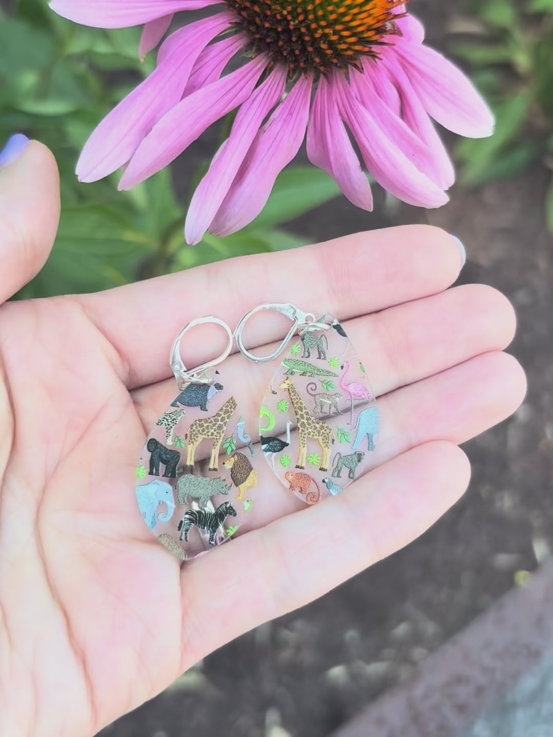Video showing teardrop shaped acrylic earrings with a zoo animal pattern, including representations of various animals such as a giraffe, lion, zebra, and others, displayed on a hand with a grassy background with a pink flower. 