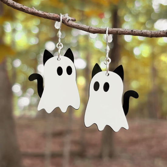 A pair of earrings hanging from a tree branch in the woods shaped like cat ghosts with eerie music. 