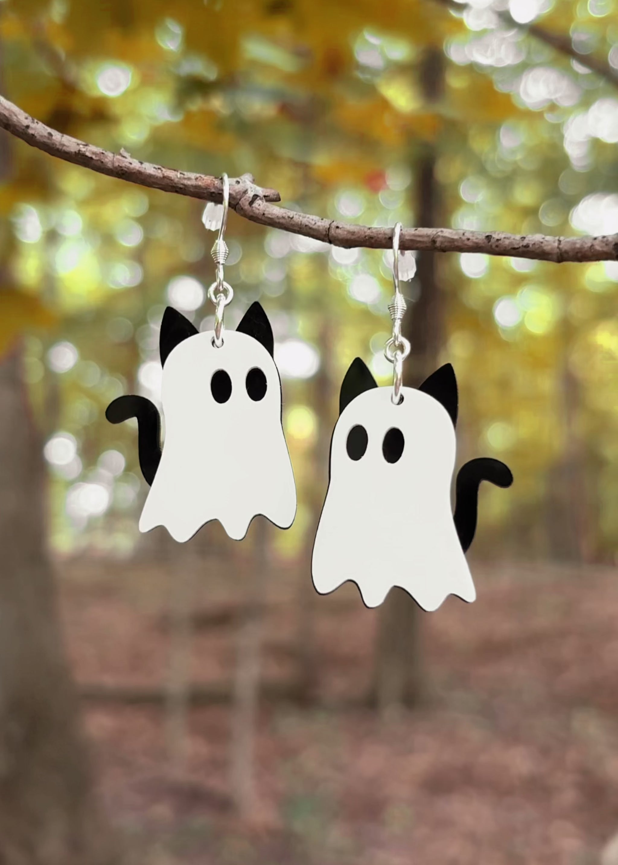 A pair of earrings hanging from a tree branch in the woods shaped like cat ghosts with eerie music. 